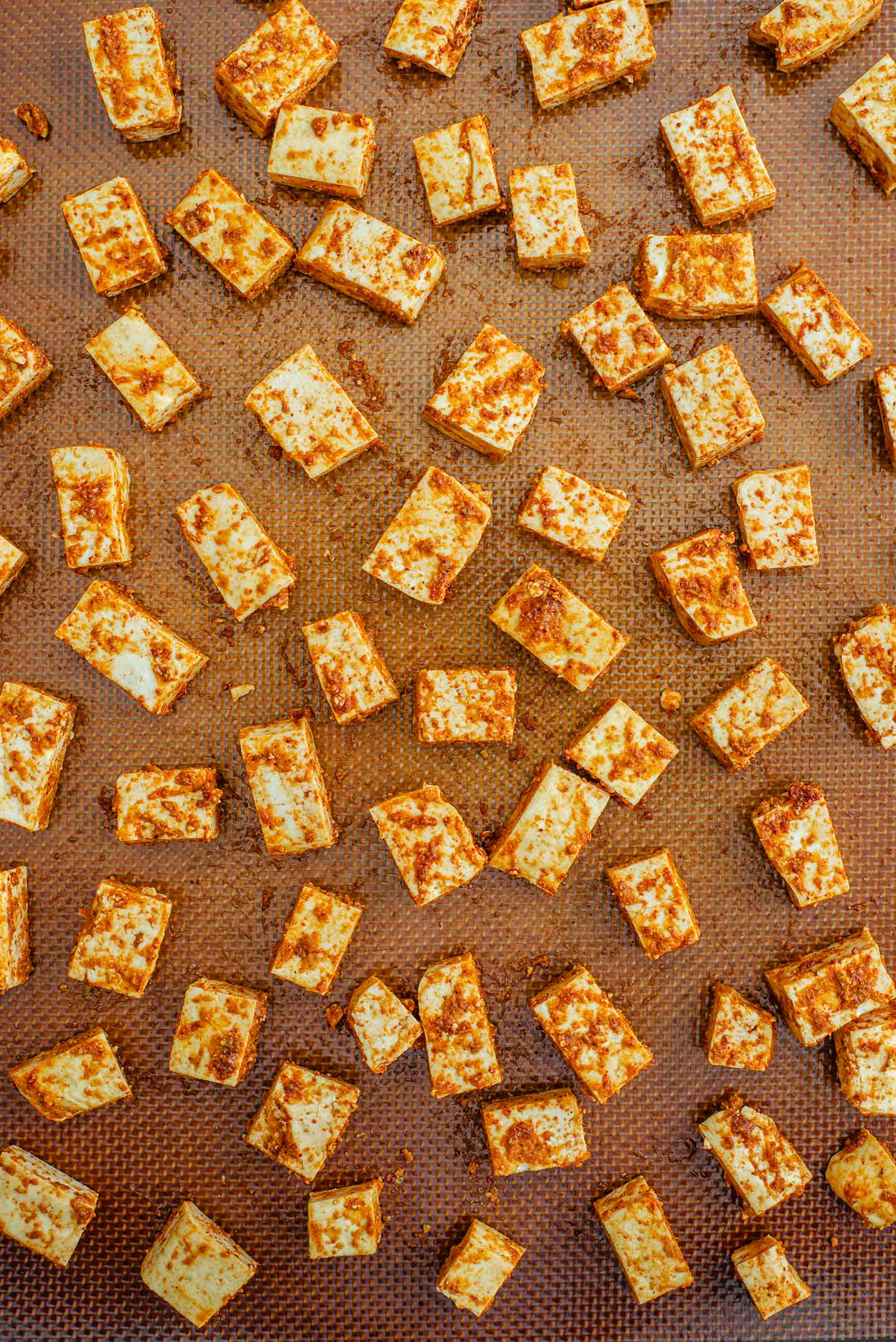 Top down view of seasoned tofu cubes spread on a baking sheet lined with a silicone baking mat prior to being baked.