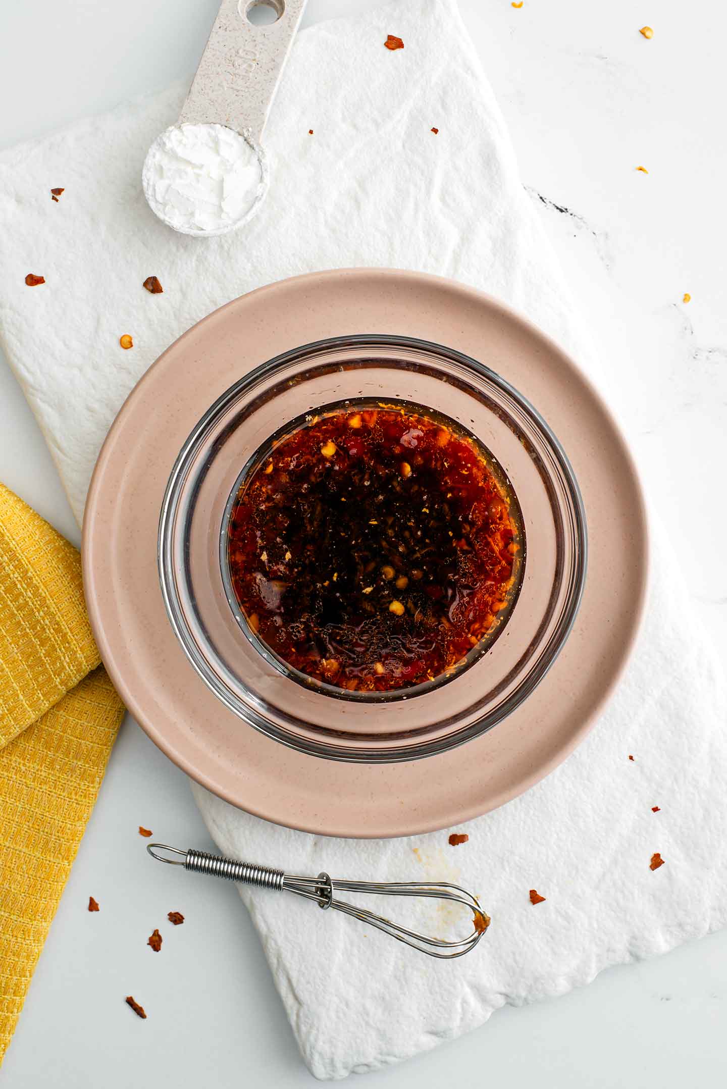 Top down view of a dark stir fry sauce in a small glass bowl. The sauce is speckled with garlic, ginger and chili flakes. Beside the sauce is a measuring spoon with starch and a small whisk.