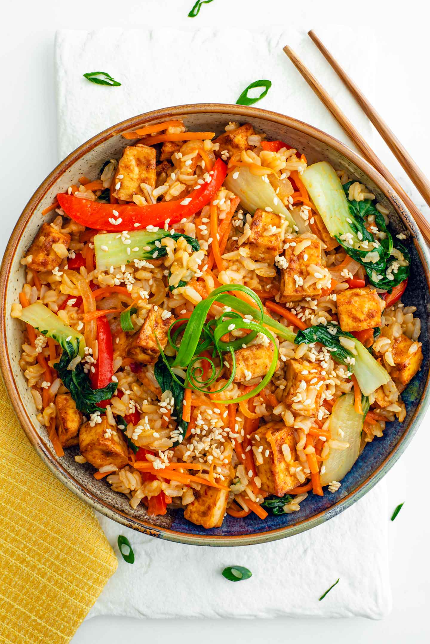 Top down view of my stir fry recipe with crispy baked tofu. Stir fried rice with red pepper, bok choy, sliced carrot and crispy tofu is garnished with curled green onion and toasted sesame seeds. 