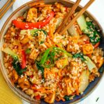 Top down view of a bowl of stir fry with crispy tofu. Chopsticks rest in the stir fried rice and vegetables garnished with curled green onion and toasted sesame seeds.