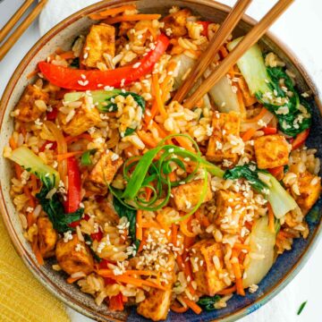 Top down view of a bowl of stir fry with crispy tofu. Chopsticks rest in the stir fried rice and vegetables garnished with curled green onion and toasted sesame seeds.