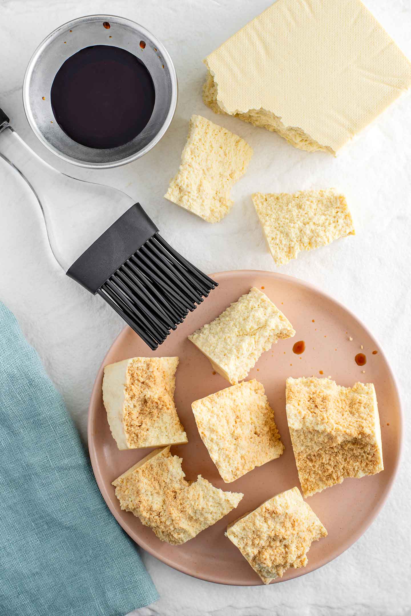 Top down view of frozen tofu torn into chunks to create small nuggets. Some nuggets are brushed with tamari using a pastry brush.