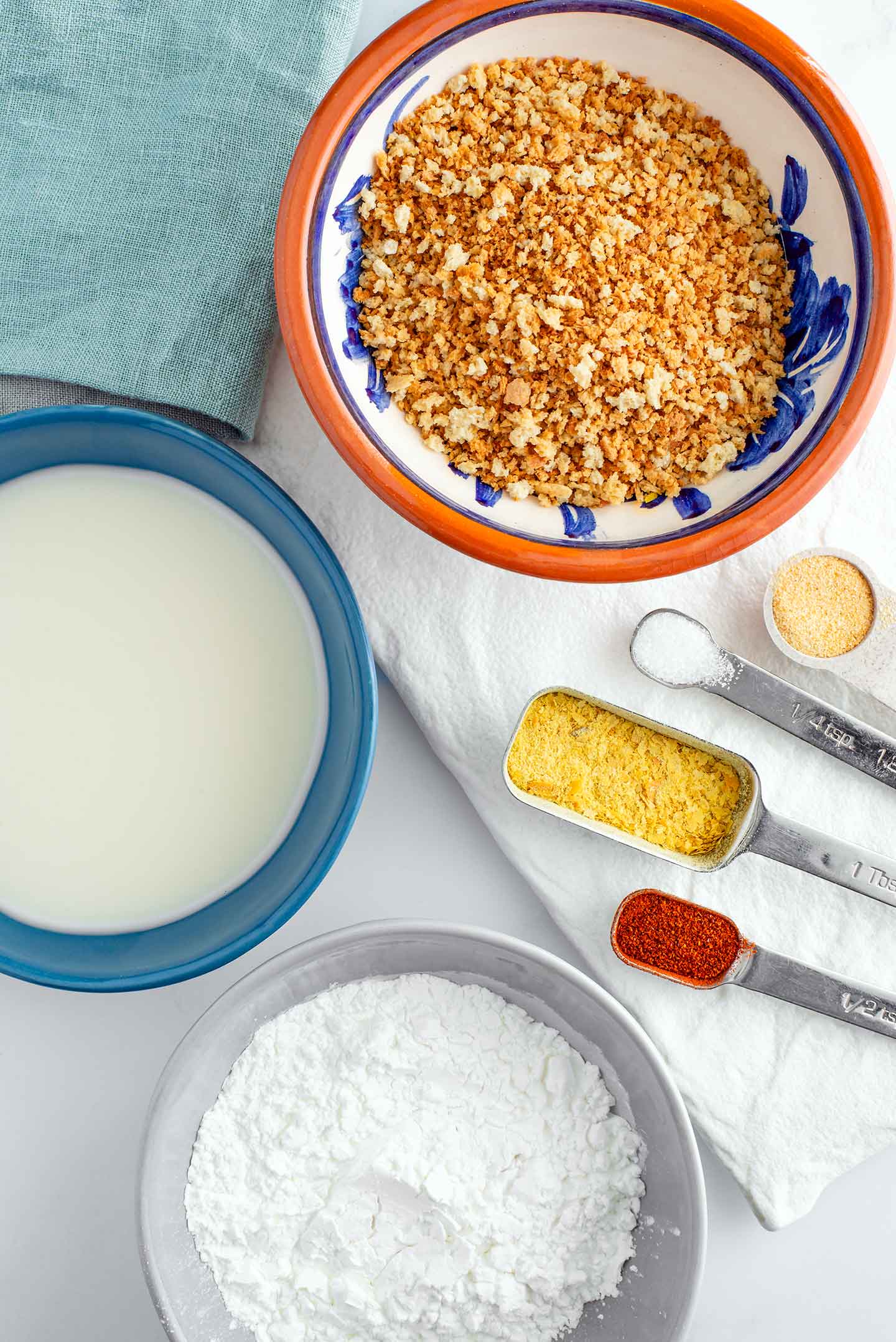 Top down view of three small dishes and some seasonings. One dish holds toasted breadcrumbs, another holds vegan buttermilk, and the third holds cornstarch.