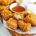 Close up of baked tofu nuggets on a tray with a dipping sauce. The white tofu centre of one golden and crispy nugget is displayed.