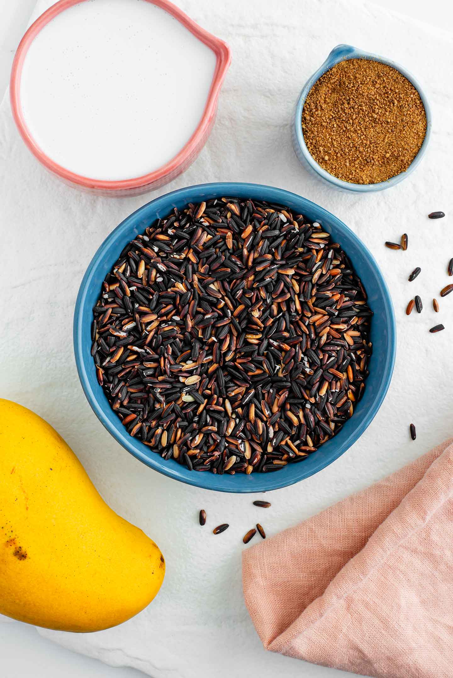 Top down view of ingredients on a white tray. Black glutinous rice fills a small bowl while coconut milk, coconut sugar and an ataulfo mango surround.