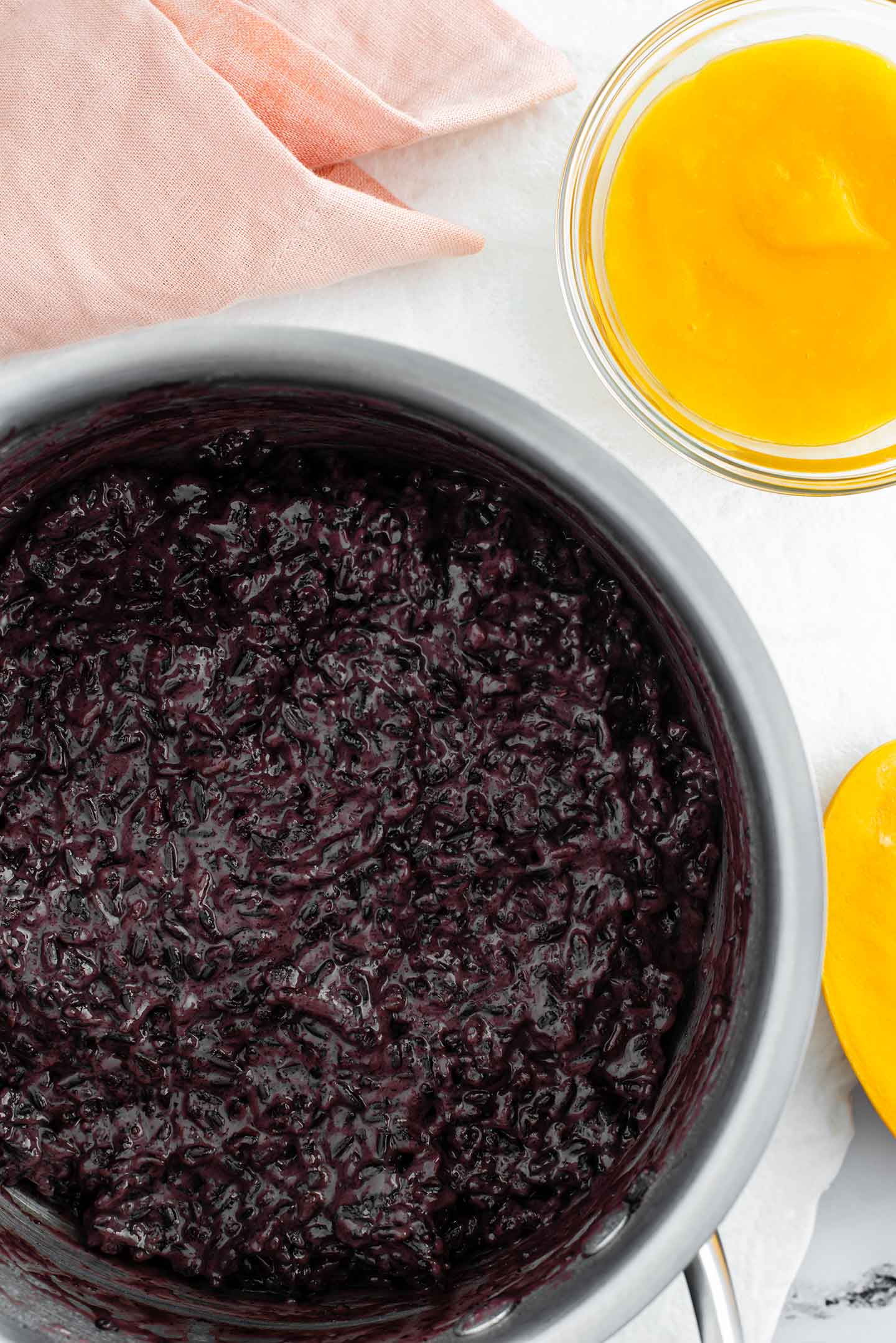 Top down view of cooked black rice pudding in a pot. Puréed mango fills a small glass bowl next to the pot.