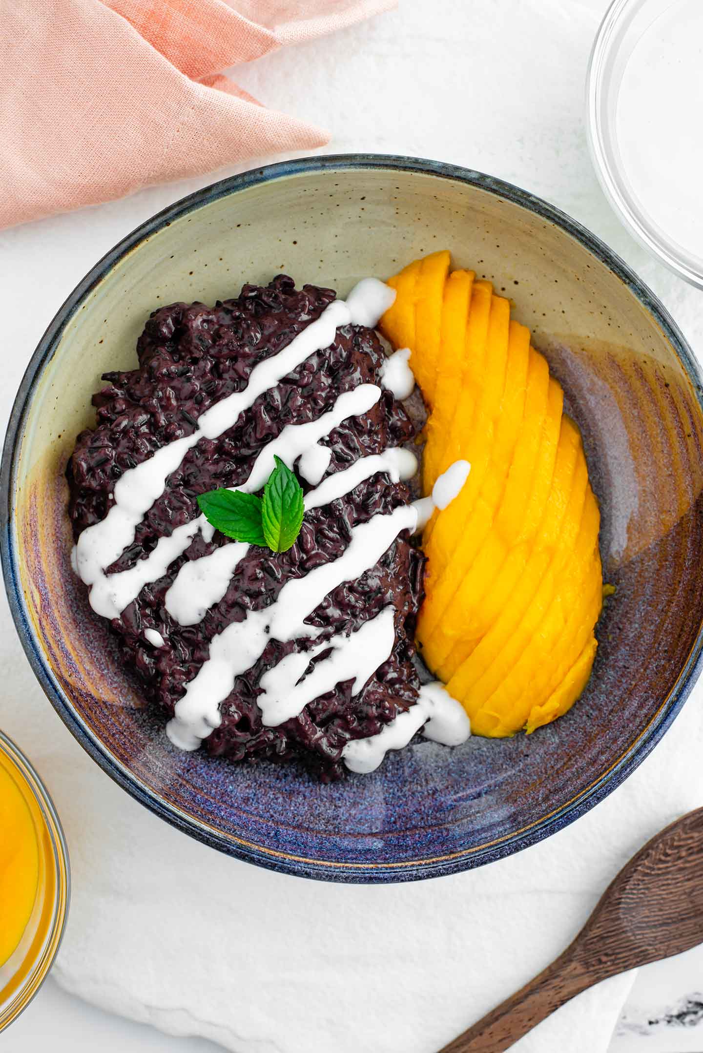 Top down view of black rice pudding in a shallow bowl with half a sliced mango to the side. A coconut drizzle decorates the top of the sticky rice and mint leaves garnish.