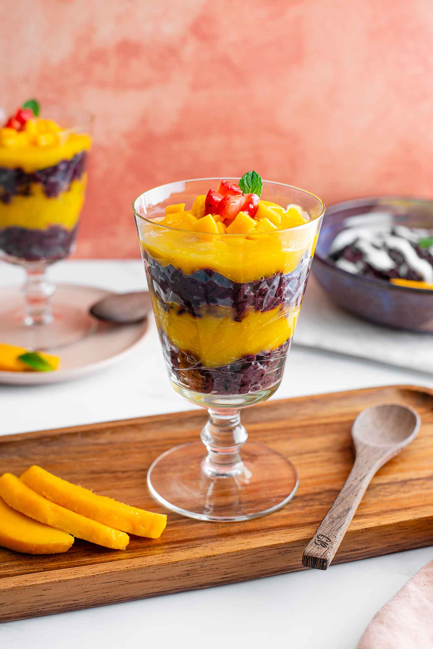 Side view of a black sticky rice parfait in a beautiful glass. Layers of pureéd mango contrast the black rice pudding and the parfait is garnished with cubed mango, strawberry, and a mint leaf. Another parfait fills the background.