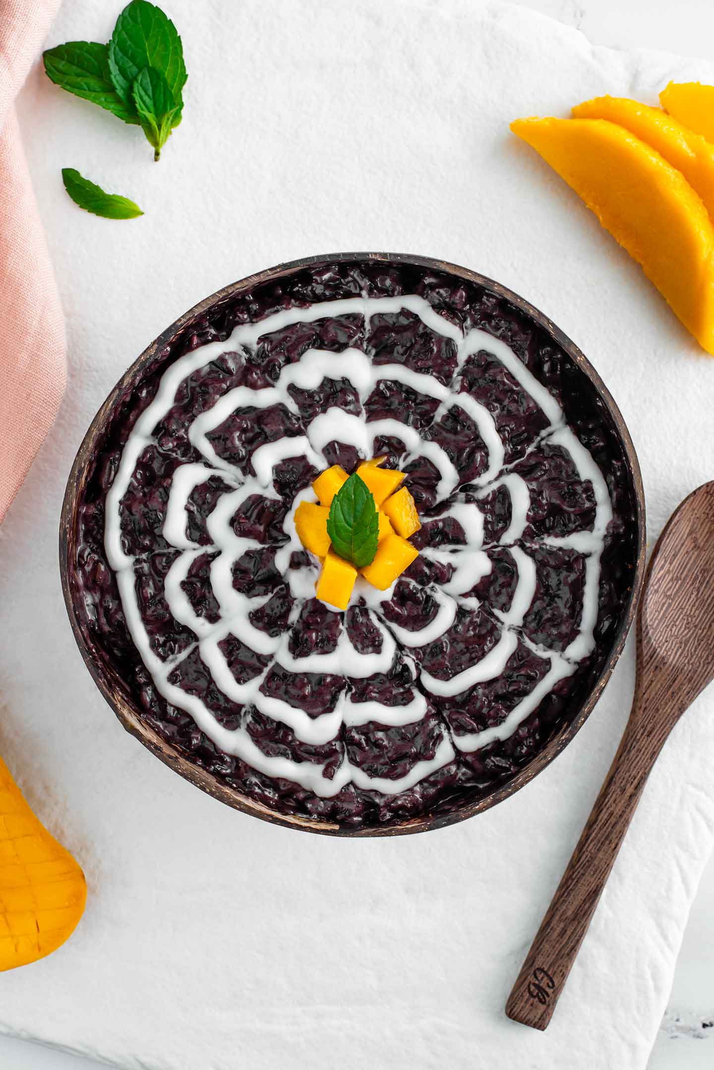 Top down view of black rice pudding in a bowl made from a hollowed out coconut. A white coconut drizzle makes a decorative marbled design on top of the black rice. Cubed mango garnishes the centre of the bowl with a mint leaf. 