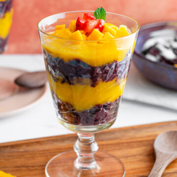 Side view of a coconut black sticky rice parfait. Layers of pureéd mango contrast the black rice pudding and the parfait is garnished with cubed mango, strawberry, and a mint leaf.