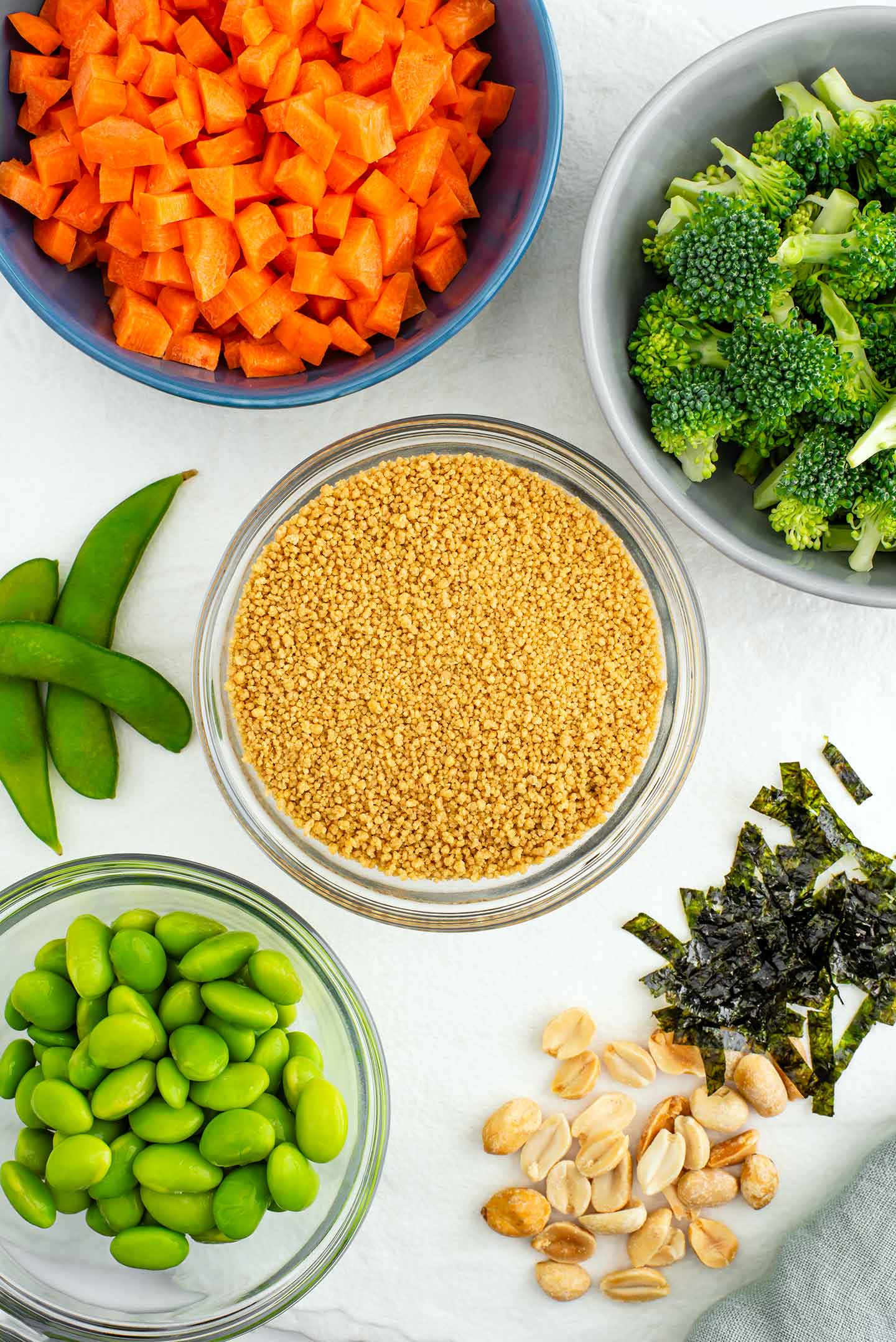 Top down view of couscous in a small bowl surrounded by bowls of broccoli, carrots, and edamame. Chopped roasted peanuts and sliced seaweed sheets fill out the remaining space on the tray.