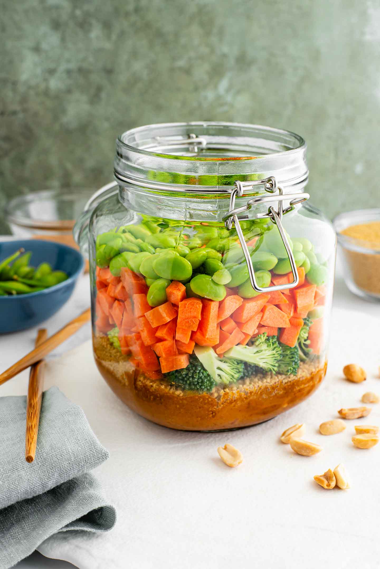 A large glass jar is filled with a layered couscous salad. The bottom layer is a peanut dressing with couscous on top. Then broccoli, carrots, and edamame. Surrounding the jar are roasted peanuts and other ingredients from the salad.