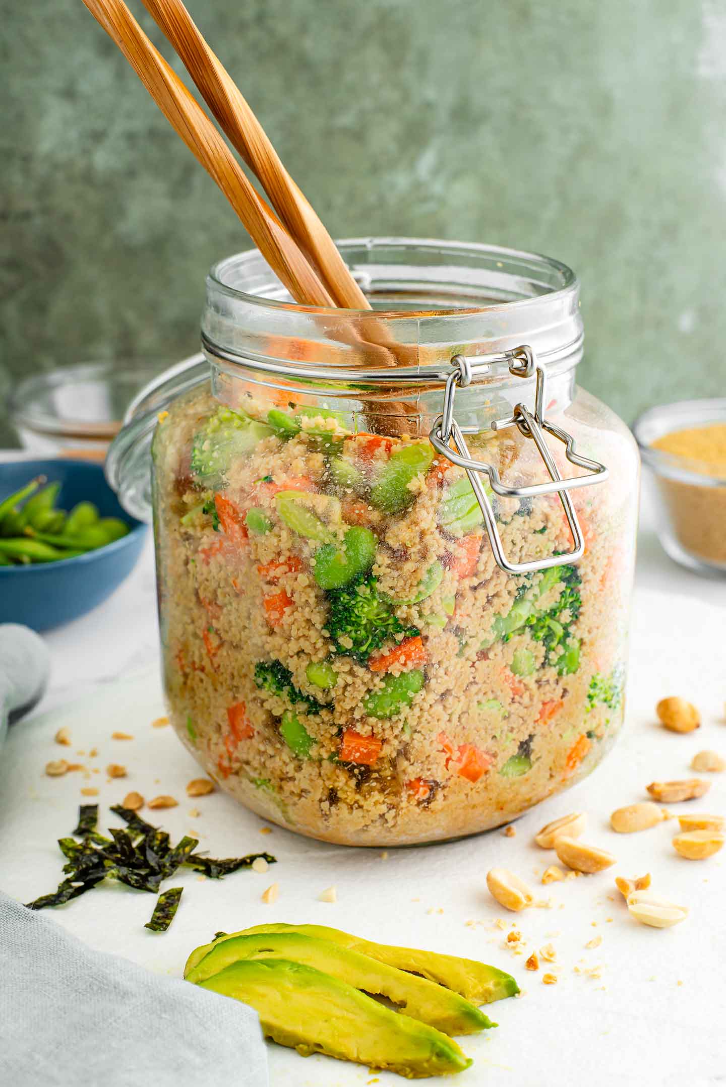 A stirred couscous pour-over salad fills a large glass jar. Chopsticks protrude from the top of the salad filled with broccoli, carrots, edamame and couscous covered in a peanut dressing.