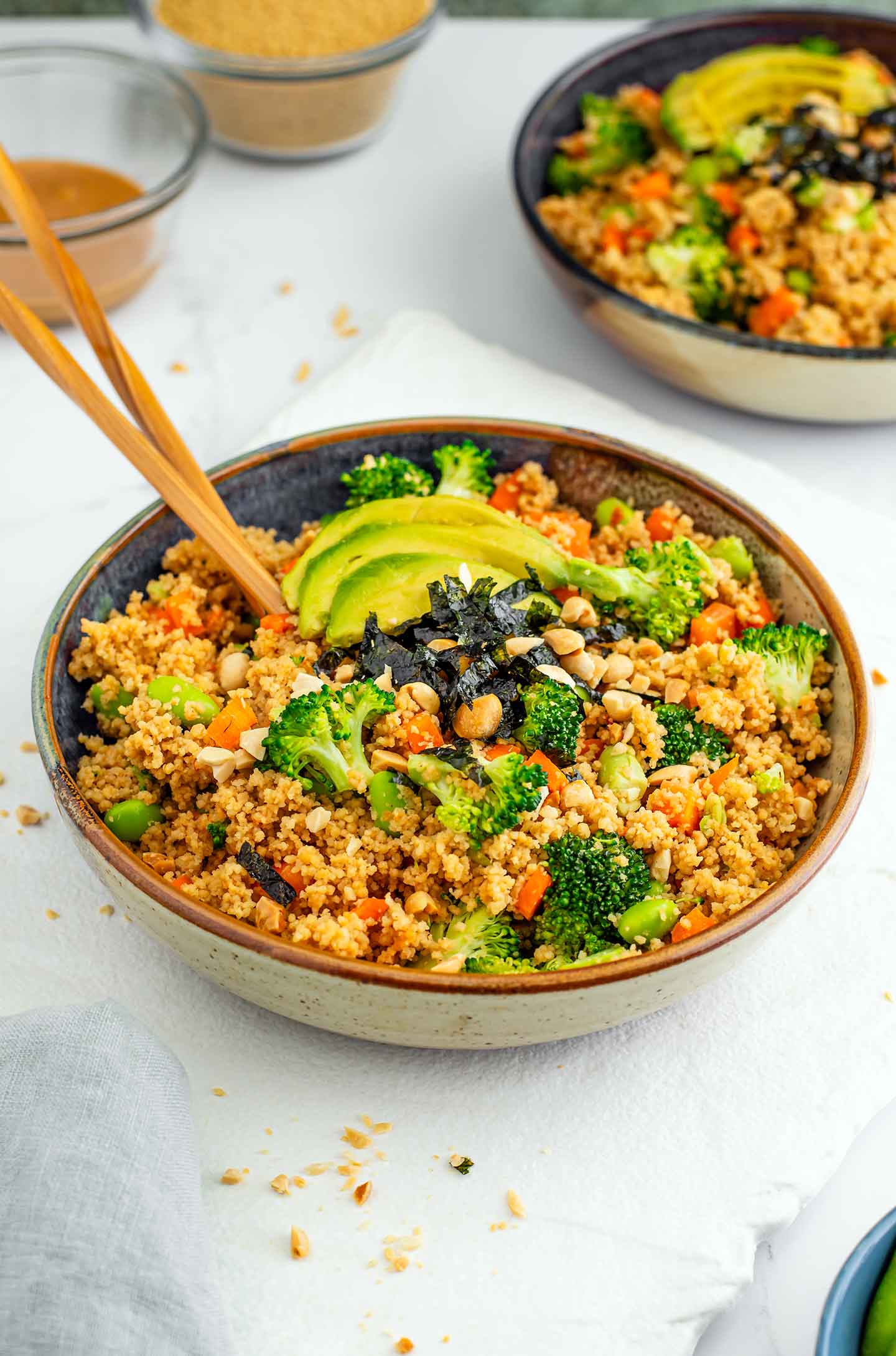 Top down view of a couscous salad in a serving bowl. The salad is topped with sliced avocado and seaweed snack sheets. Chopsticks rest in the colourful salad filled with broccoli, carrots, and edamame.