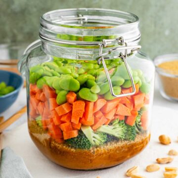 A large glass jar is filled with a layered couscous salad. The bottom layer is a peanut dressing with couscous on top. Then broccoli, carrots, and edamame.