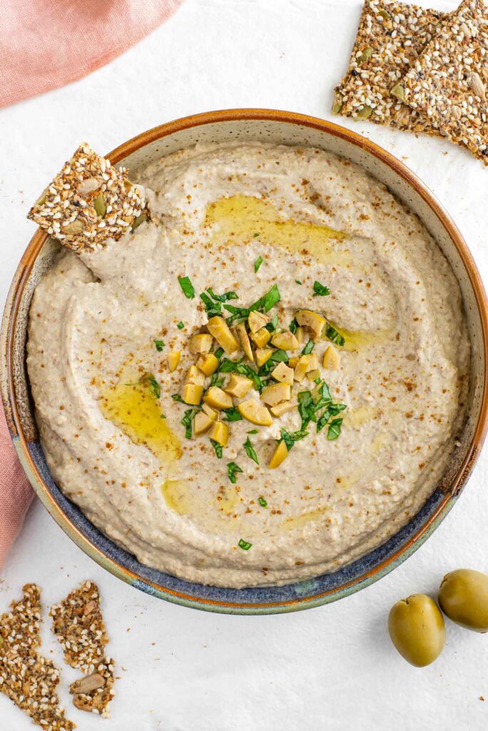 Top down view of creamy green lentil hummus in a shallow bowl. The centre is garnished with chopped olives and basil. Seed crackers and Spanish olives surround the bowl.