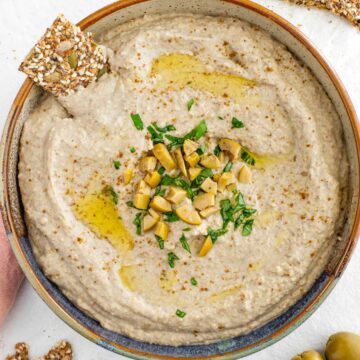 Top down view of creamy green lentil hummus in a shallow bowl. The centre is garnished with chopped olives and basil. A seed cracker rests in the hummus.