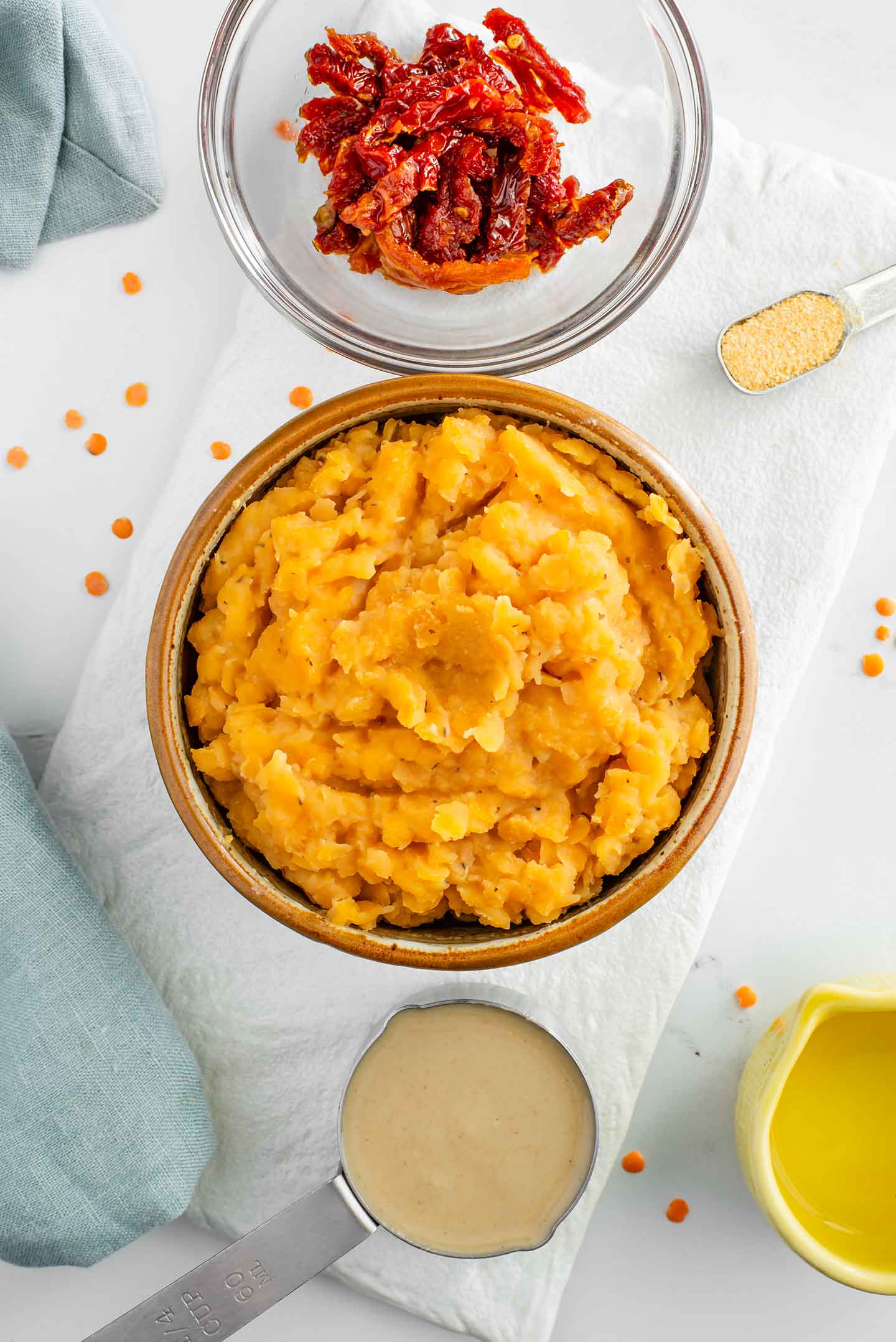 Top down view of ingredients on a white tray. A bowl with cooked red lentils surrounded by sun-dried tomatoes, tahini, lemon juice, and garlic powder.