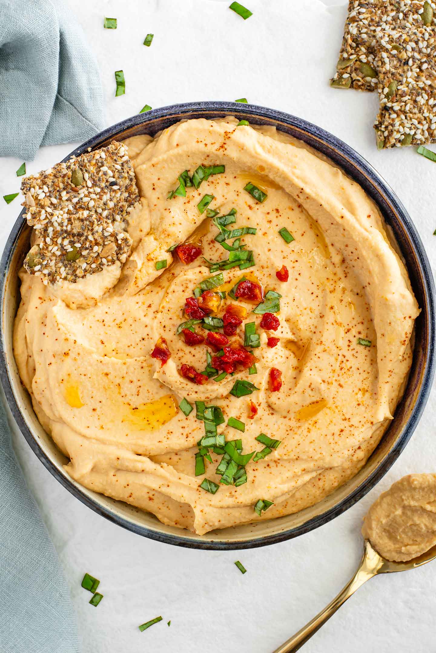 Top down view of a shallow bowl filled with creamy red lentil hummus. The centre is garnished with sun-dried tomatoes and basil. A seed cracker rests in the hummus and a spoon of thick hummus rests beside the bowl.