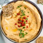 Top down view of a shallow bowl filled with creamy red lentil hummus. The centre is garnished with sun-dried tomatoes and basil. A seed cracker rests in the hummus.