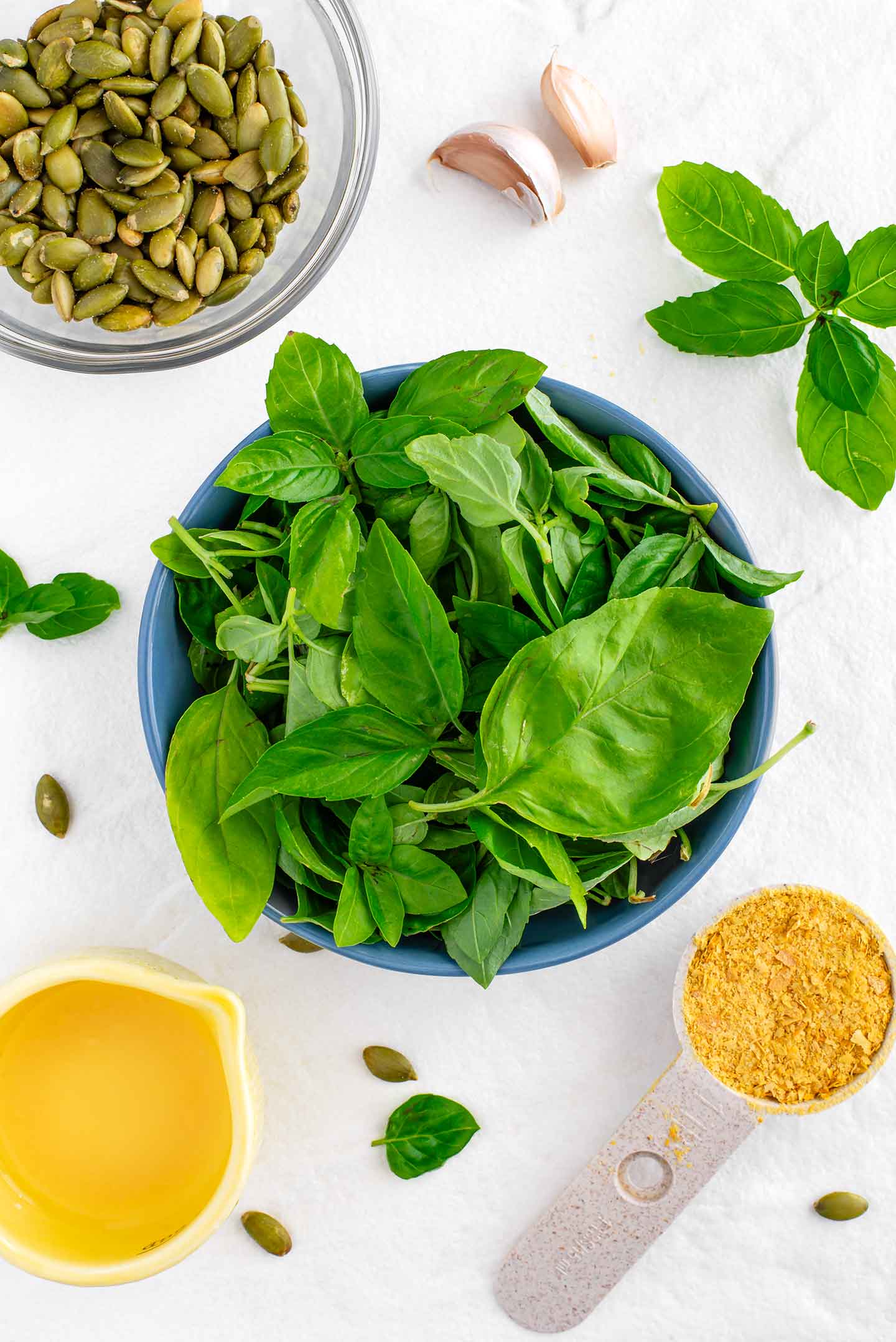 Top down view of a small bowl filled with bright green basil leaves. Pumpkin seeds, garlic cloves, lemon juice, and a tablespoon of nutritional yeast surround the basil.