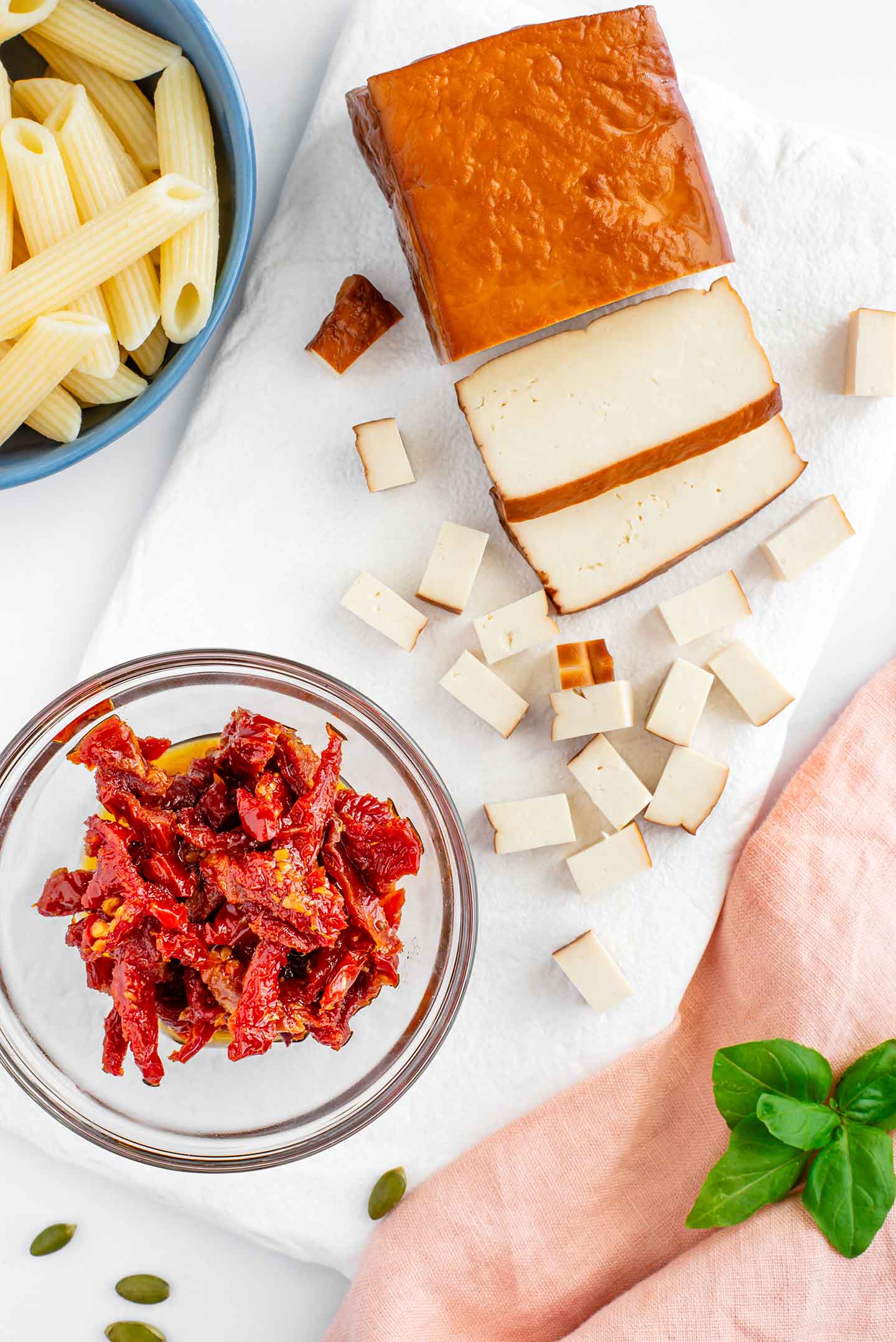 Top down view of ingredients on a white tray. A block of smoked tofu is partially diced. A small bowl contains sliced sun-dried tomatoes, and cooked penne noodles can be seen on the edge of the image. 