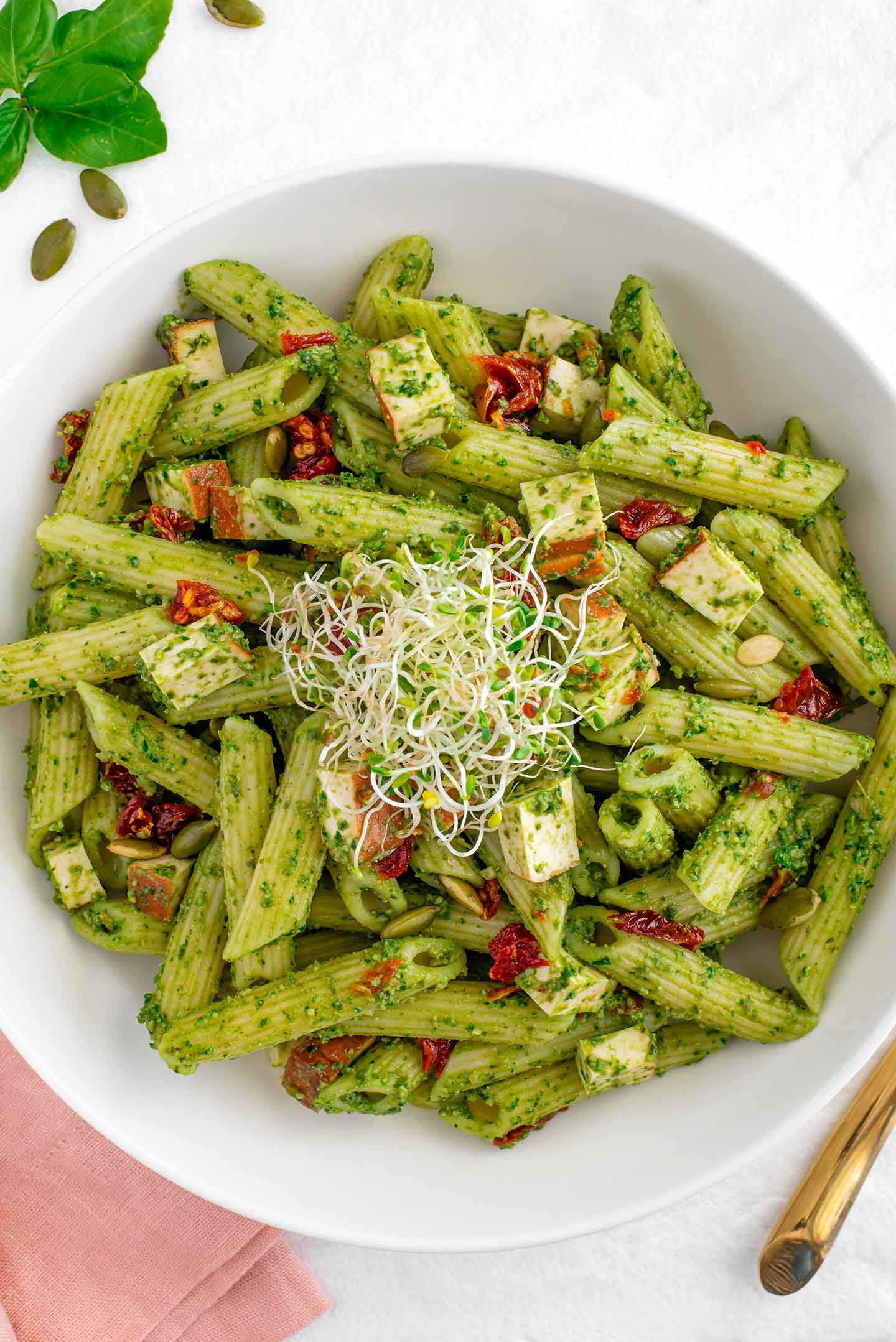 Top down view of a bowl of vegan pesto pasta. The penne noodles are coated in the bright green pesto. Sun-dried tomatoes and smoked tofu are scattered throughout. Fresh alfalfa sprouts garnish the top. 