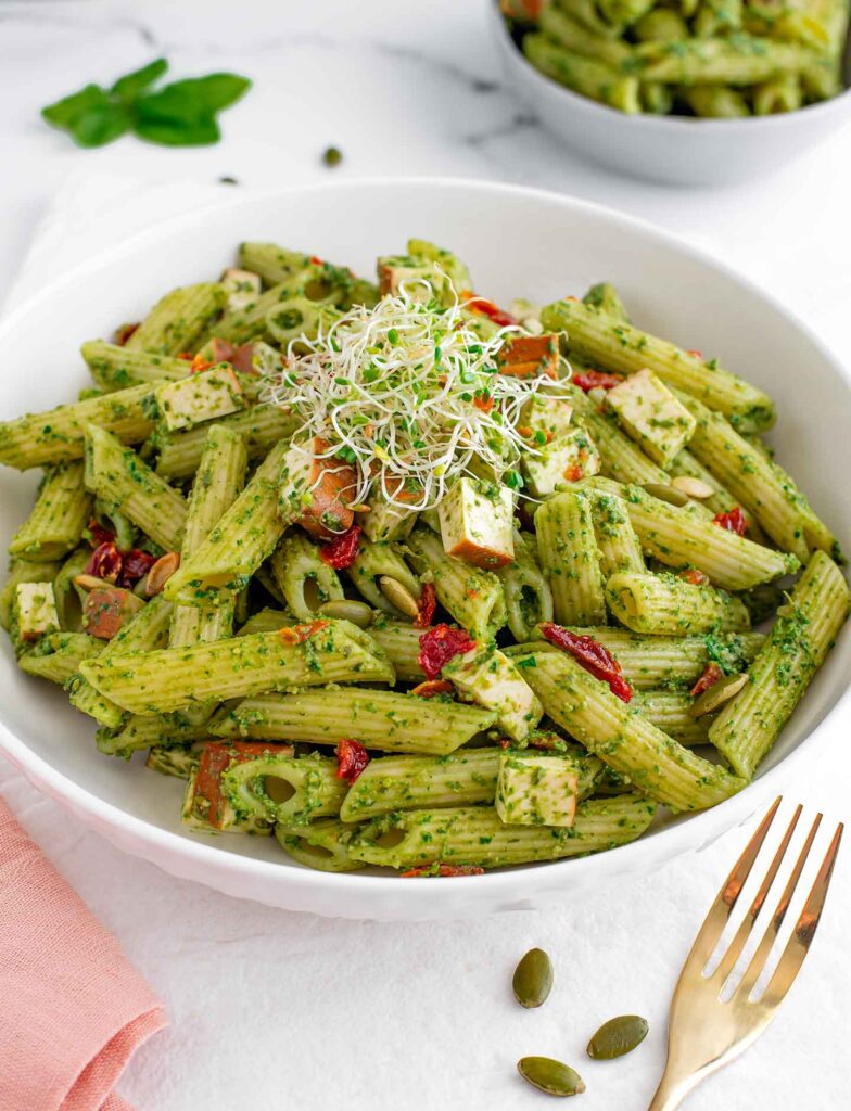 Side view of a bowl of penne noodles covered in a fresh green pesto sauce. Chopped sun-dried tomatoes and smoked tofu are scattered through the pasta. Alfalfa sprouts and pumpkin seeds garnish the top.