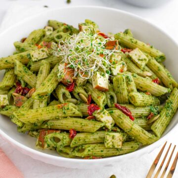 Side view of a bowl of penne noodles covered in a fresh green pesto sauce. Chopped sun-dried tomatoes and smoked tofu are scattered through the pasta. Alfalfa sprouts garnish the top.