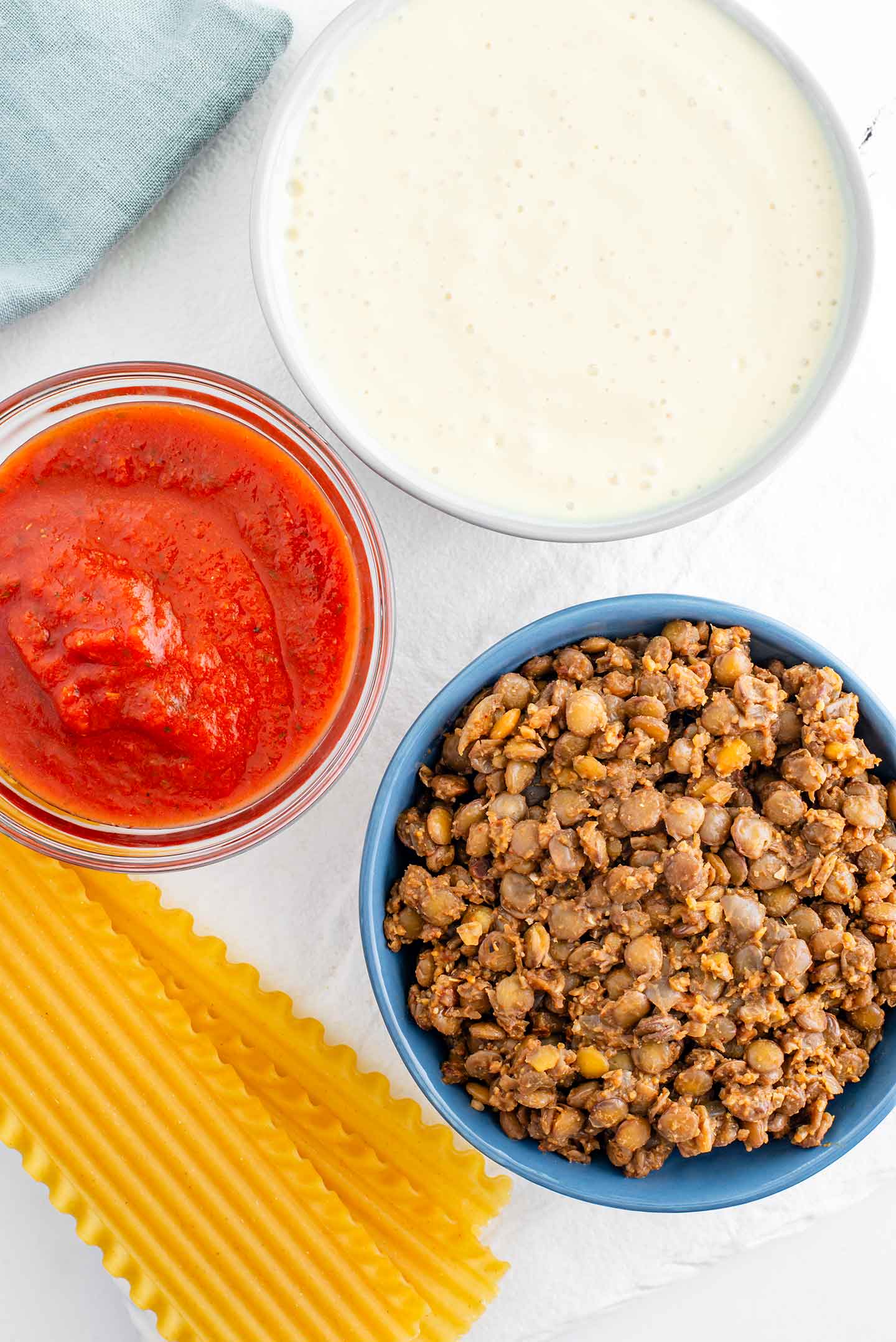 Top down view of the components of a lasagna. Savoury lentil meat sauce, a blended tofu cheese sauce, tomato sauce, and dry lasagna sheets. 