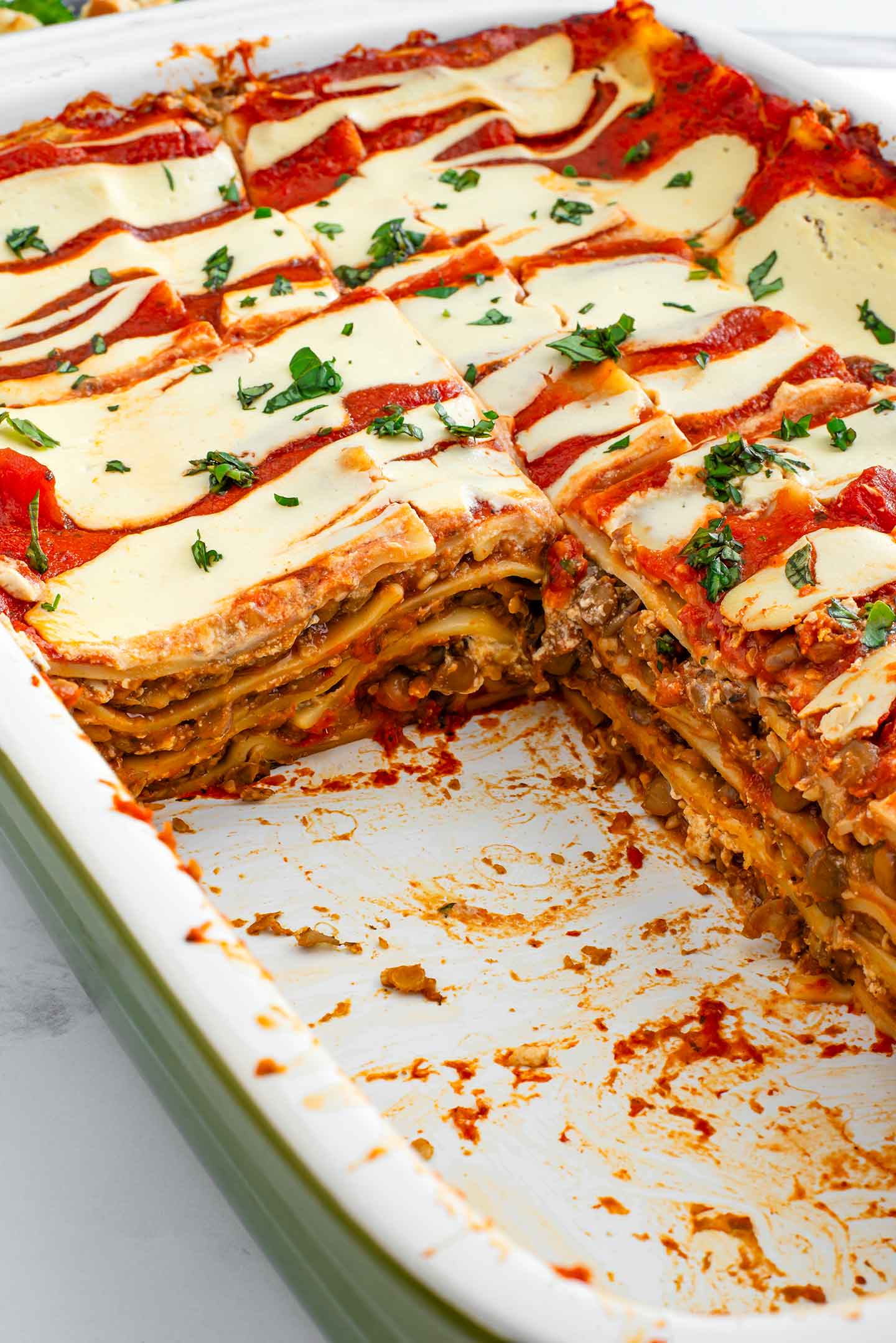 Two slices are missing from a vegan lentil lasagna in a casserole dish. The layers of pasta, lentil meat, tomato sauce, and tofu cheese are clearly defined.