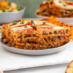 A slice of vegan lentil lasagna rests on a plate. The lasagna is full of a moist lentil meat and a creamy tofu cheese sauce.