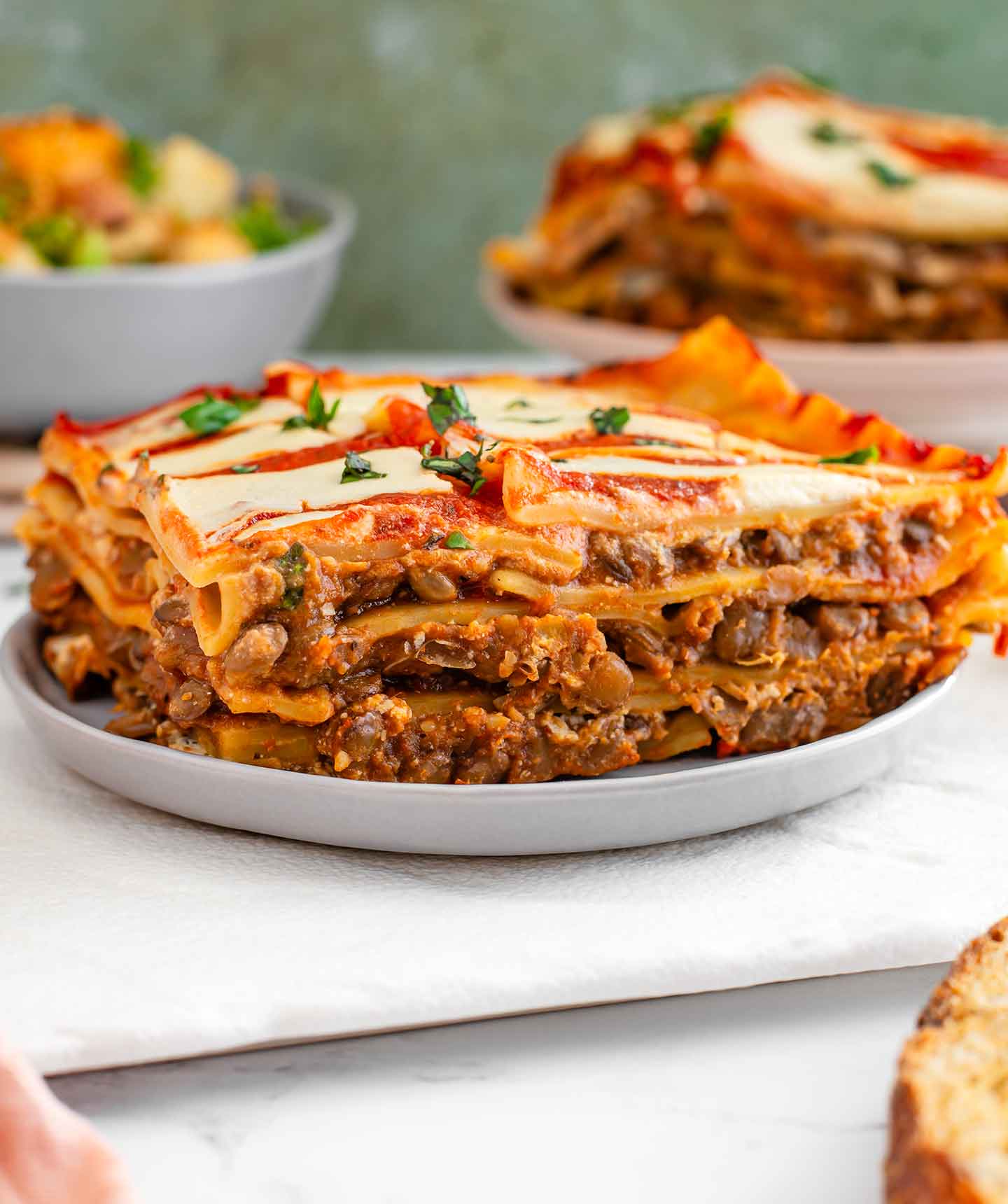 A slice of vegan lentil lasagna rests on a plate. The lasagna is full of a moist lentil meat and a creamy tofu cheese sauce. Chopped basil garnishes the top of the slice.