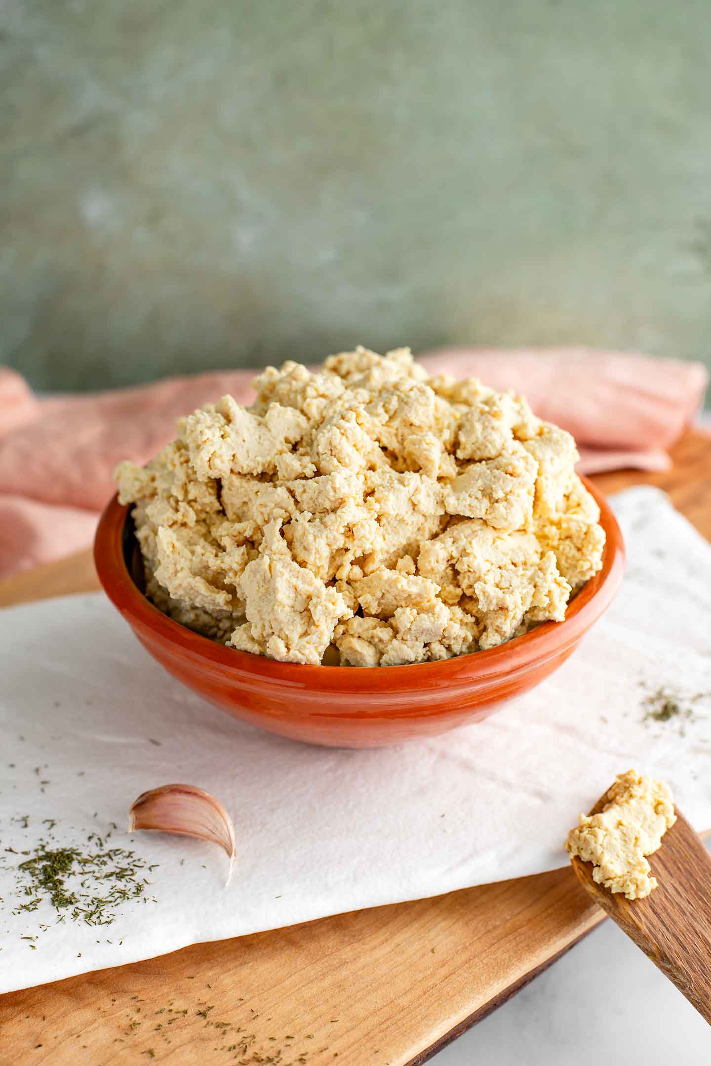 Side view of a crumby but moist tofu ricotta in a small bowl. Dried dill and a garlic clove garnish a tray beneath the cheese substitute.