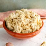 Close-up side view of a crumby but moist tofu ricotta in a small bowl. Dried dill and a garlic clove garnish a tray beneath the cheese substitute.