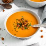 Side view of bright orange, creamy soup. The soup is thick and topped with crispy roasted chickpeas.