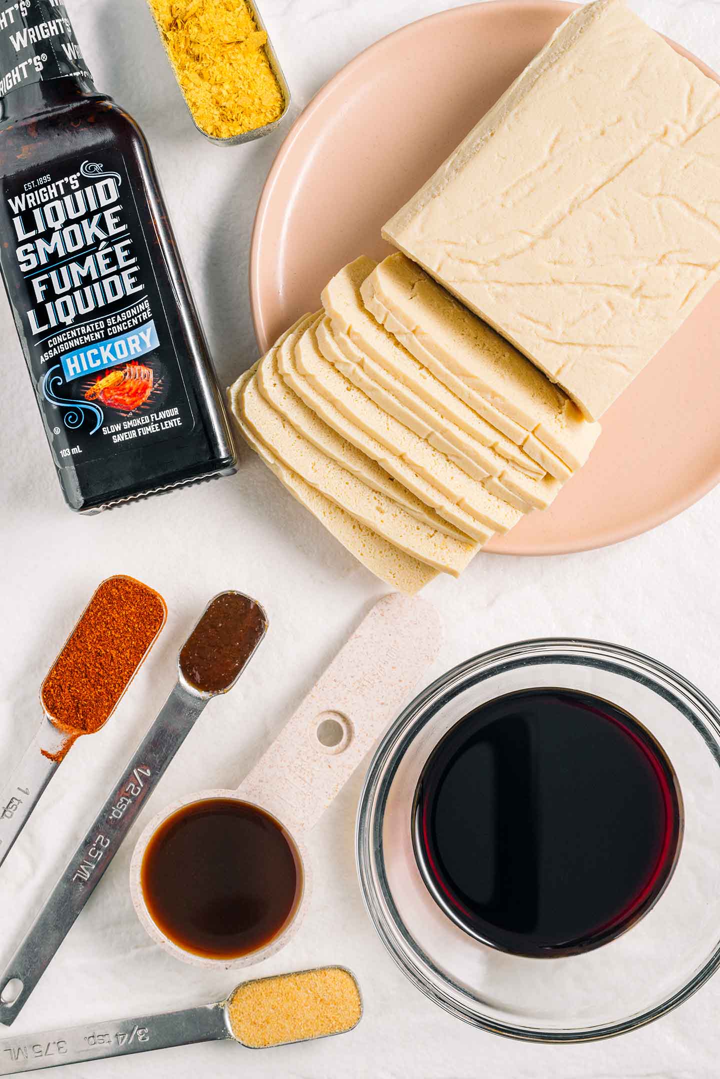 A block of previously frozen firm tofu is being sliced thinly and the texture is obviously spongey. Tamari, worcestershire sauce, liquid smoke, and other seasonings surround the tofu.