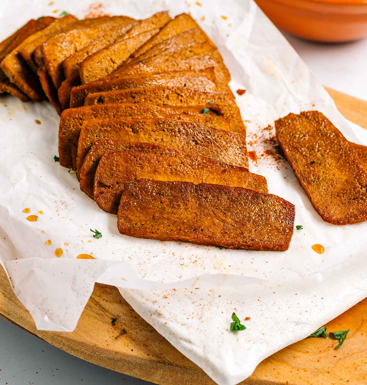 Slices of golden and juicy tofu shaved meat are laid out on a piece of parchment paper on top of a wooden tray. The tofu meat looks textured and spiced.