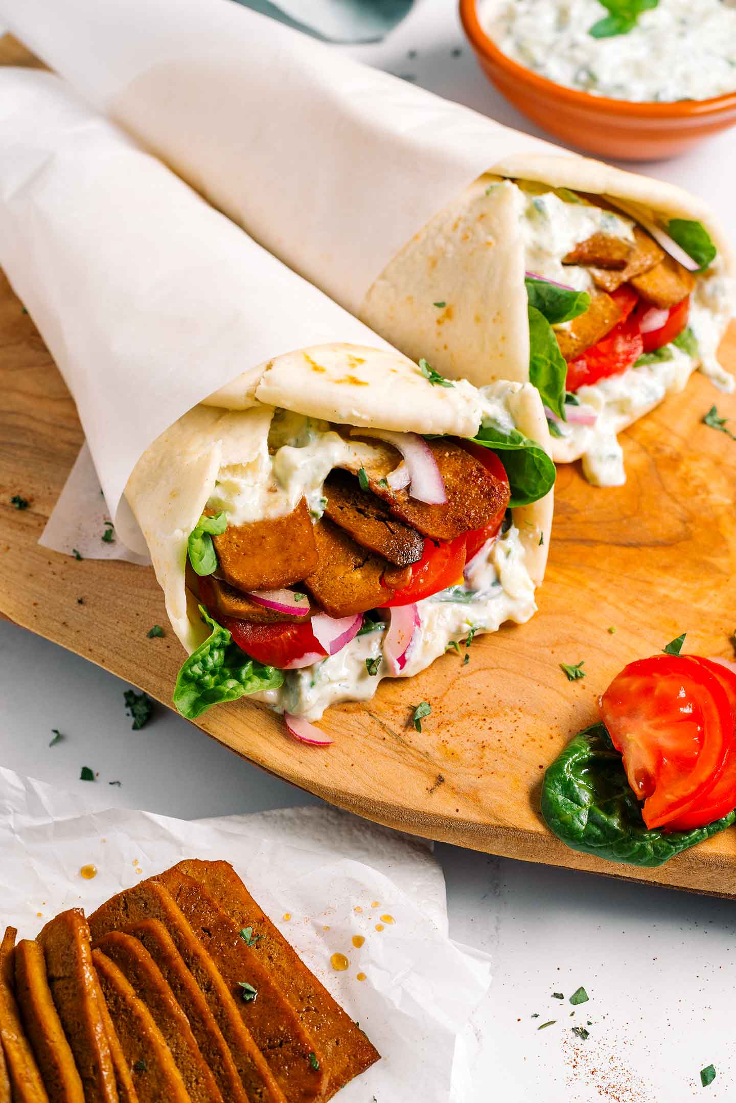 Top down view of two vegan gyro on a tray wrapped in fluffy pita bread and filled with vegan tzatziki, tofu shaved meat, lettuce, red onion, and tomato.