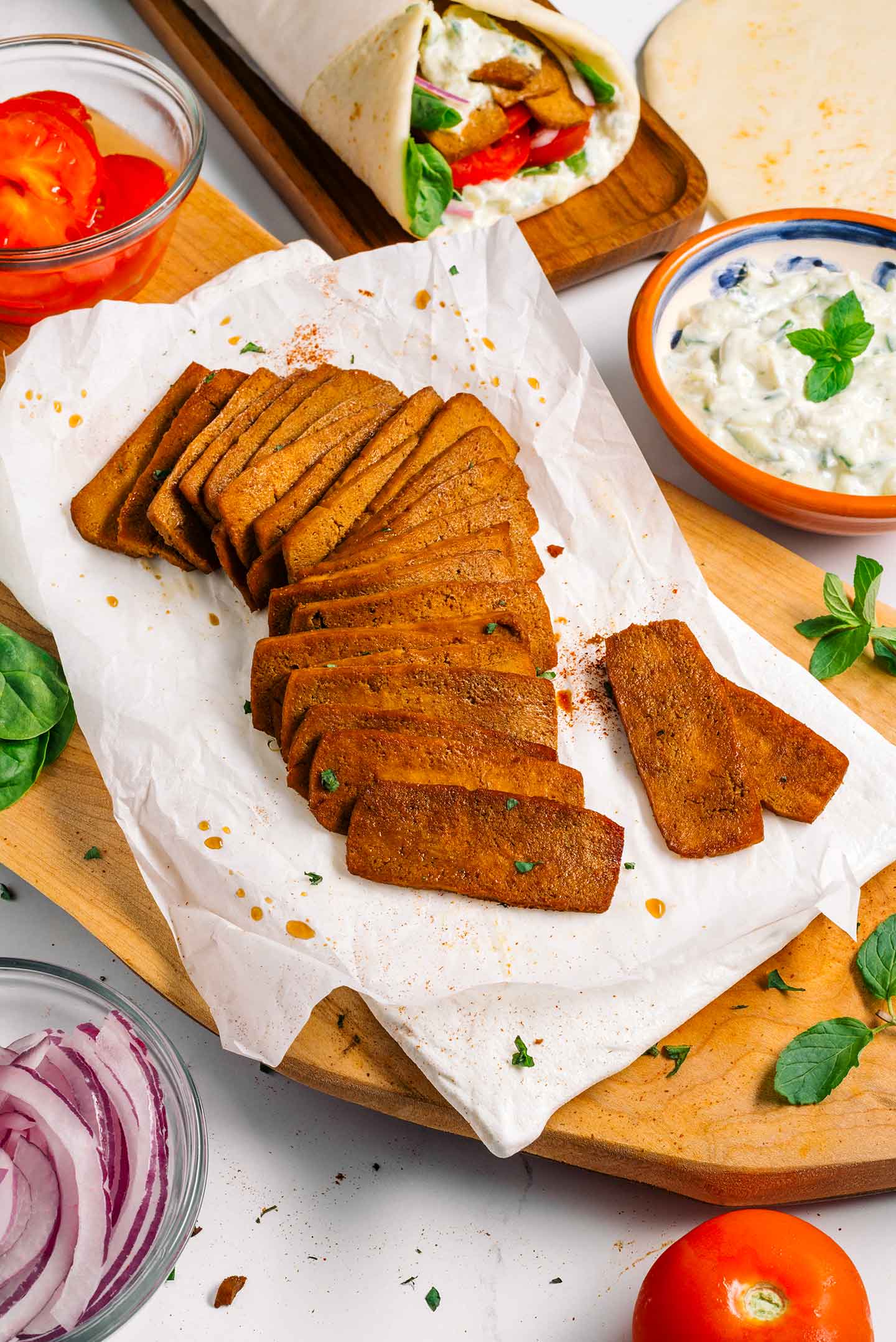 Top down view of tofu ingredients for vegan gyro. Slices of tofu shaved meat, a bowl of creamy tzatziki, lettuce, tomato, onion, and Greek pita bread.