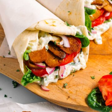 A fluffy Greek pita is filled with creamy tzatziki sauce, lettuce, tomato, red onion, and juicy tofu shaved meat.