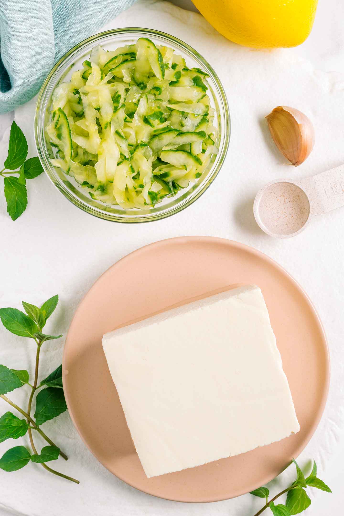 Top down view of ingredients. A block of soft tofu, a bowl of grated cucumber and a clove of garlic. Plus some vinegar, lemon, and sprigs of mint.