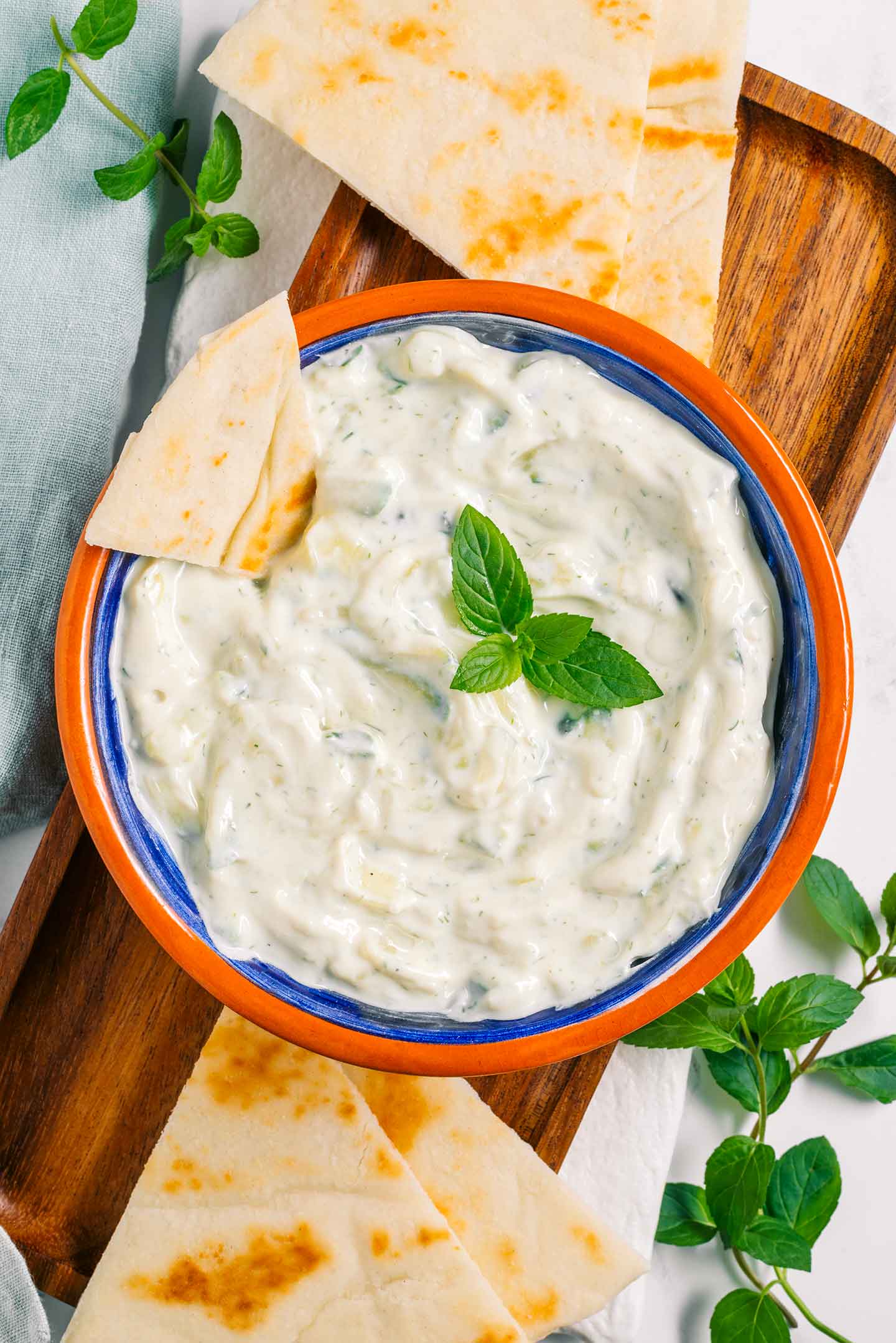 A creamy vegan tzatziki sauce speckled with cucumber and fresh herbs fills a small bowl. Slices of pita bread and sprigs of herbs surround the creamy dip.