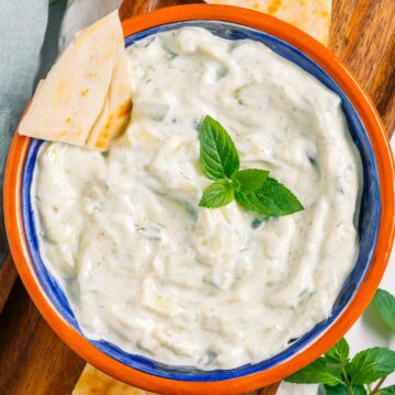 A creamy vegan tzatziki sauce speckled with cucumber and herbs fills a small bowl. A slice of pita bread rests in the dip and fresh herbs garnish the top.