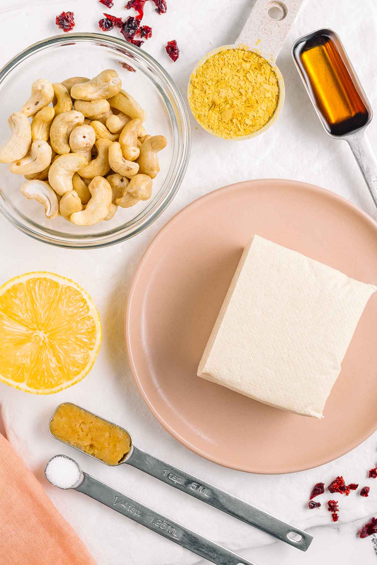 Top down view of tofu, cashews, a halved lemon, nutritional yeast, maple syrup, miso paste, salt, and dried cranberries.