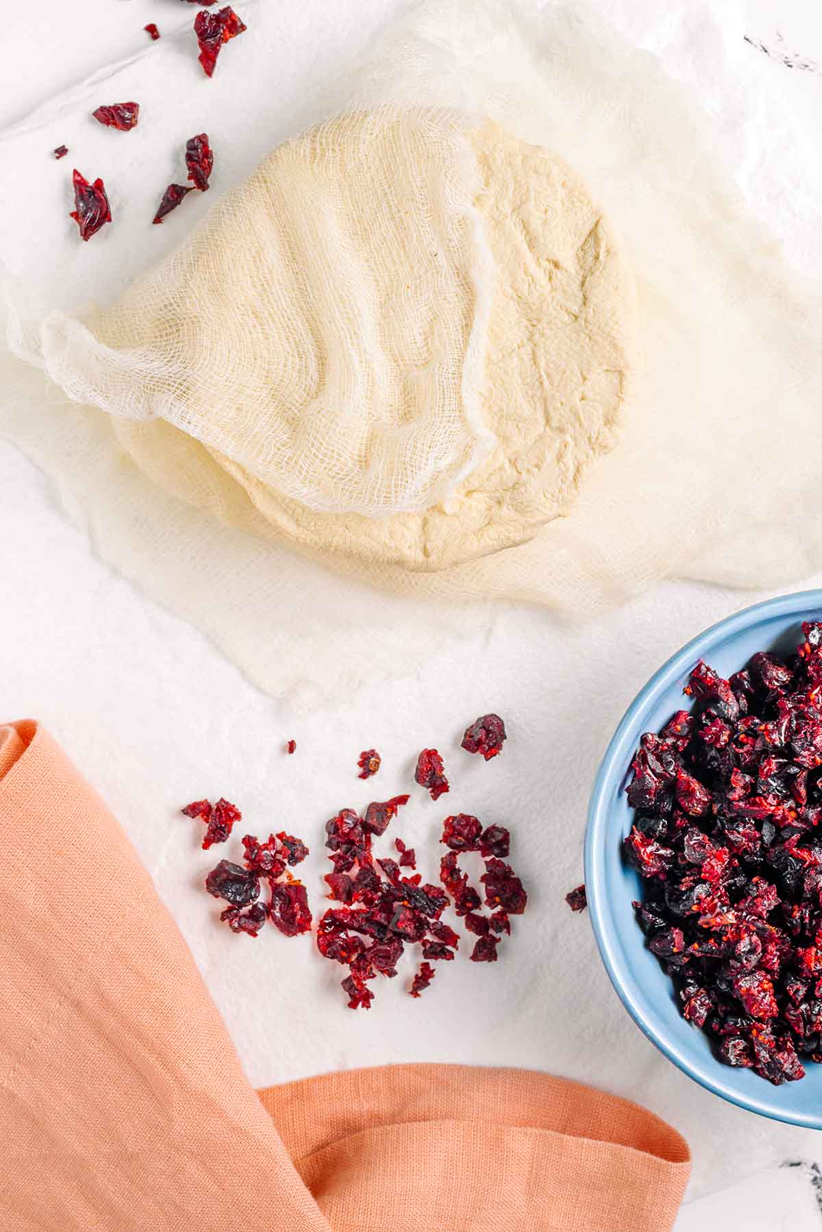 Top down view of a vegan cheese ball wrapped in cheesecloth. A bowl of chopped dried cranberries sits beside the cheese ball.