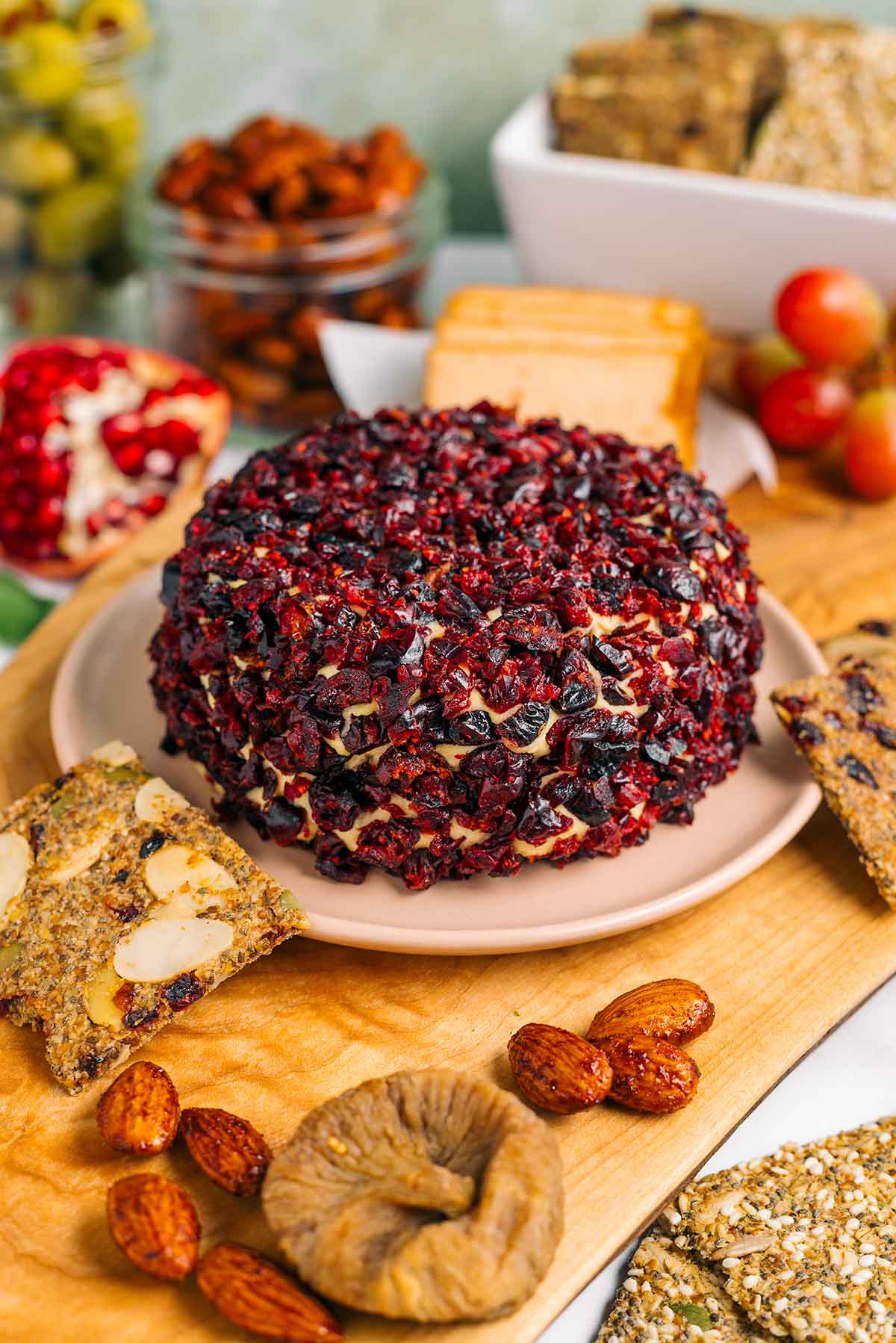 Side view of a cranberry crusted vegan cheese ball in the centre of a vegan charcuterie board. The cheese ball has not been cut. Crackers, nuts, fruits, smoked tofu and olives surround.