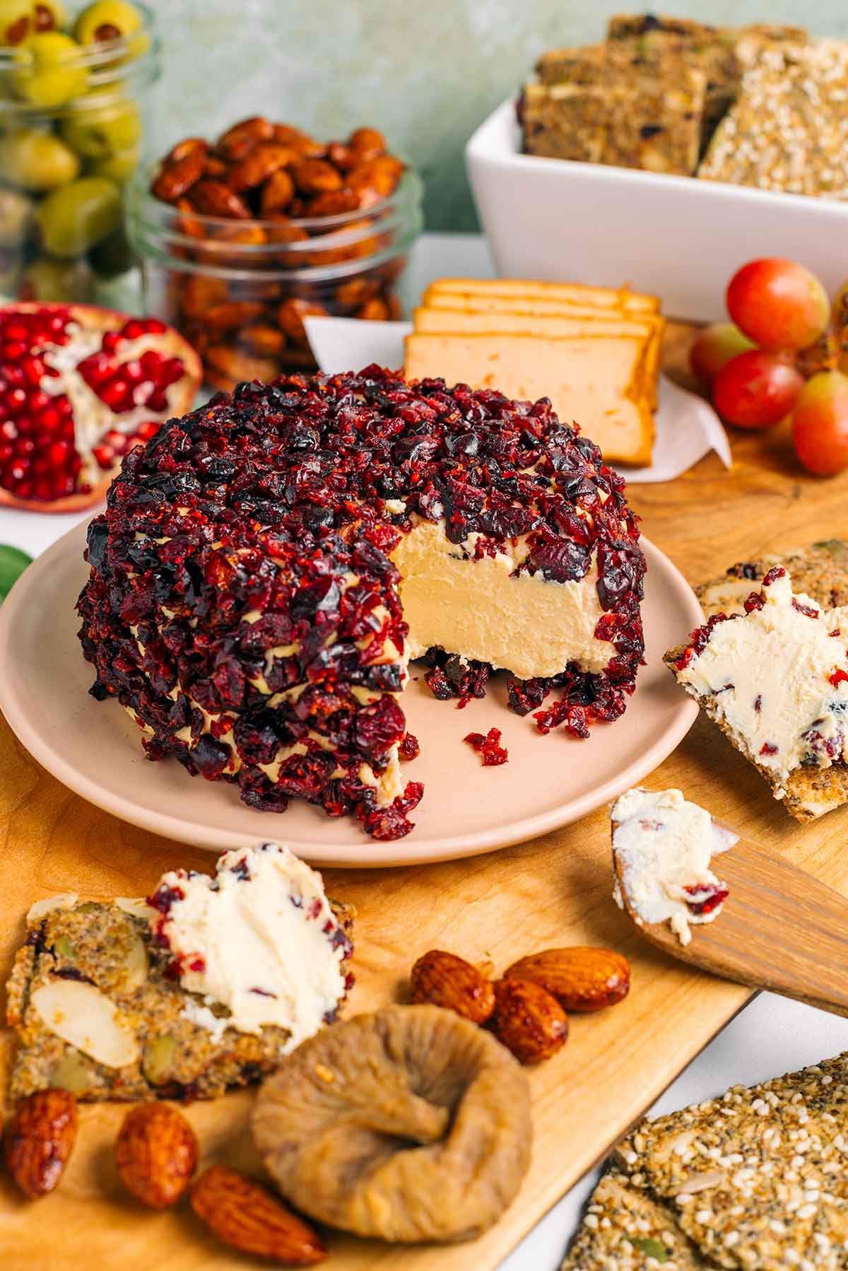 A vegan cheese ball coated in chopped dried cranberries has a wedge removed. The cheese in the centre is soft, spreadable, and garnishing crackers on a charcuterie board.