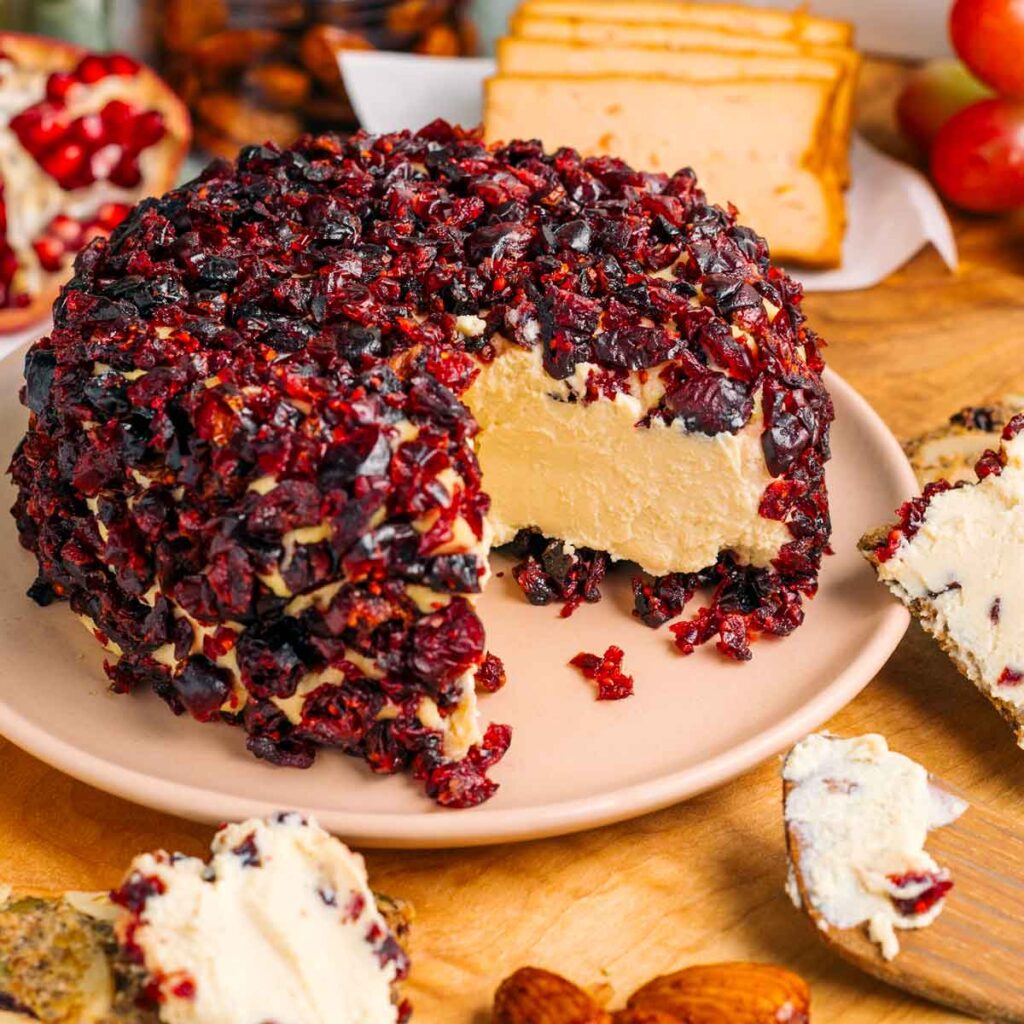 A vegan cheese ball coated in chopped dried cranberries has a wedge removed. The cheese in the centre is soft and spreadable.