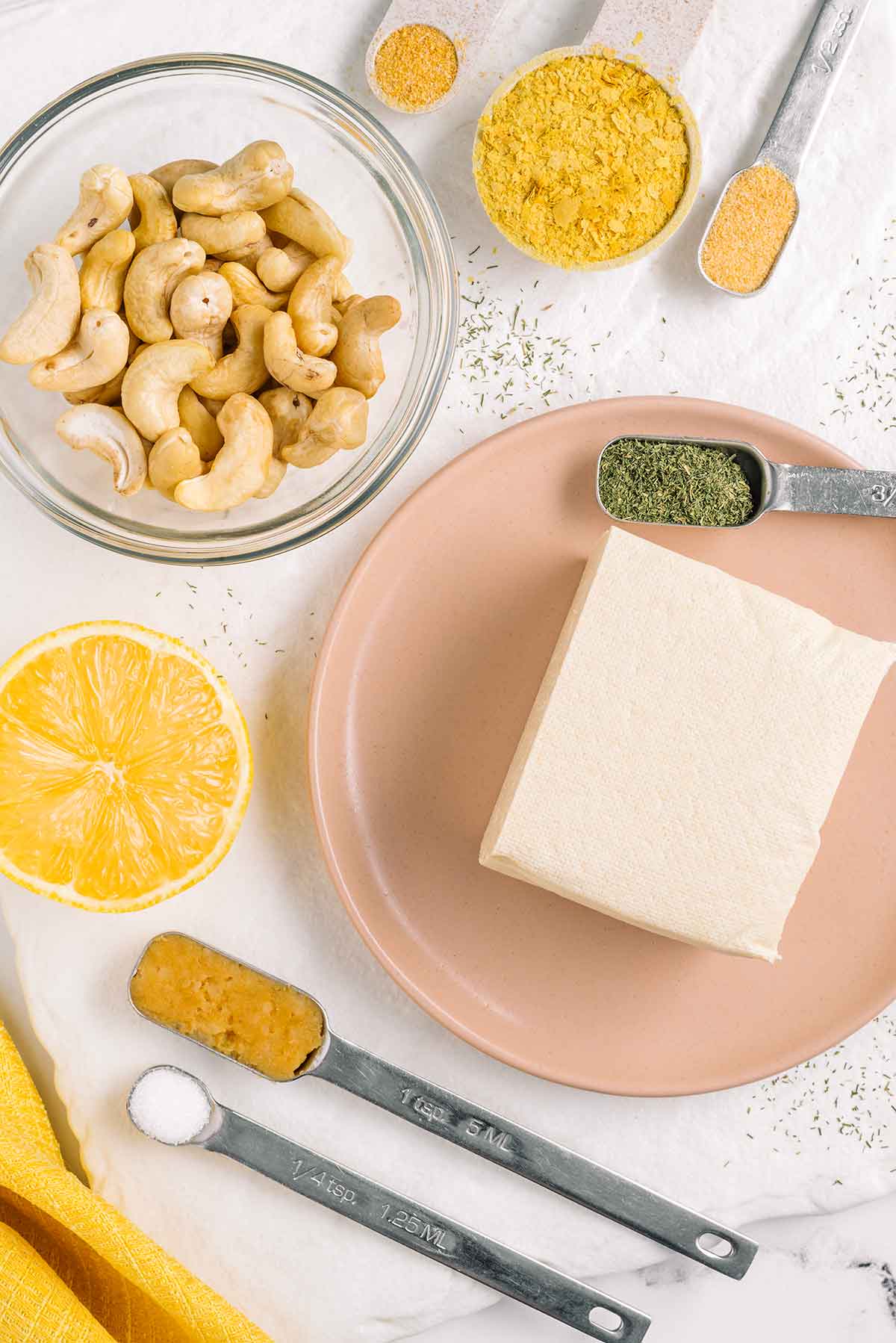 Top down view of tofu, cashews, nutritional yeast, a halved lemon, miso paste, salt, garlic powder, onion powder, and dried dill.